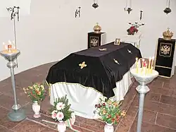 Small chamber with white walls and pink marble floor. In the middle is a tomb on three small stairs from the same marble, covered with white silk and black velvet with embroidered golden Greek crosses. It is surrounded by candles and flowers.