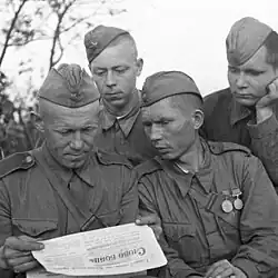 A black and white photo of a group of soldiers all reading a single newspaper.