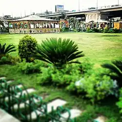 Al-Sabil Park. In the foreground is a fence, a path, and some ornamental shrubs. In the background is short grass.