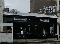 A one-story building, painted white with black trimmings, is seen in a 3/4 view surrounded by multi-story buildings.