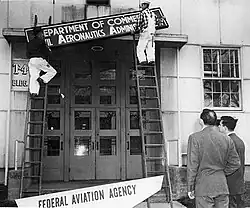 Workmen on ladders are swapping an old "Civil Aeronautics Administration" sign for a new "Federal Aviation Agency" sign above a tall doorway of a building.