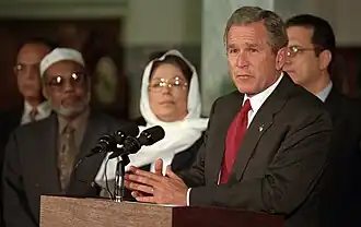 George W. Bush, president of the United States in 2001, stands at a pulpit with two microphones. He wears a black lightly pin-striped suit, a white button-up shirt, and a red necktie. An American flag pin is affixed to his lapel. Behind him, out of focus in the background, are three men and one woman. One of the men wears a kufi. The woman wears a hijab.