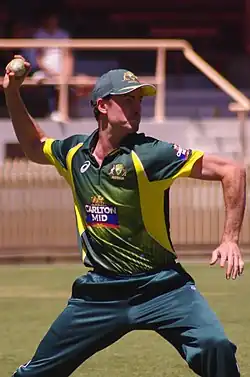 A cricketer dressed in a green uniform with a cap about to throw a cricket ball with his right hand towards the right of the photograph
