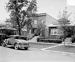 Two-story house with cars parked on the street in front of it. It is partially obstructed by a neighboring house on the right. A tall tree is on the lawn in front of the house.