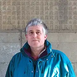 Headshot of Andew West at Cloud Platform, in front of a monument with Tangut script. He then was a middle-aged, white man with short, grey hair, wearing a large blue jacket.