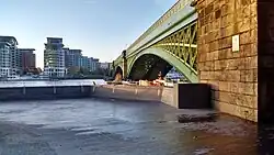 View of an empty paved area that is part of a public riverside footpath, with a railway bridge, next, to the right. Backdrop of the river Thames.