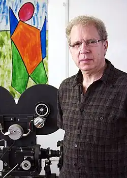 A white man with glasses, standing at side of film camera