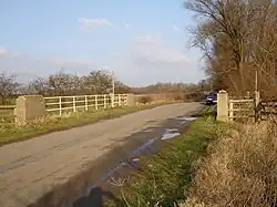 road with concrete pillars and white painted handrails where the drainage ditch passes underneath. A stand of bare trees on the right and some bare hedgerows on the left show this is winter