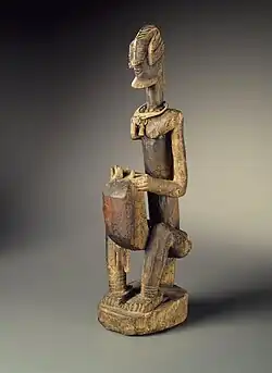 A wooden sculpture of a man seated on a low stool, playing a kora which rests on and slightly between his knees. The figure is stylized, with an elaborate hairstyle, the kora body slightly boxy.
