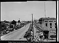 Center Street at 100 West facing west, with a parade on the south side of Center Street. Numerous automobiles line the street and businesses such as Hedquist Drugs, Wilkins Hotel, Taylor Brothers, and Smith Brothers Auto Repair Shop line the street.