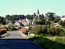 A general view of a village in tiers on the side of a wooded hill with a river in the foreground.