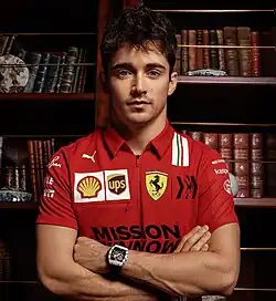 Leclerc wearing a Ferrari polo shirt and Richard Mille watch, standing with his arms folded in front of a bookshelf