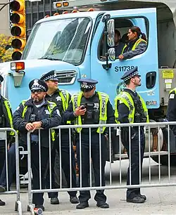A group of Chicago police officers behind a fence at a roadblock. A Chicago public works truck, also used to block the road, is behind the officers. One of the officers is looking at his cell phone.