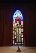 Lady Chapel stained glass window, commissioned in 2004. A wahine (Māori woman) draped in a korowai with pounamu cross necklace holding a bible, framed by Luke 1:46-47. Below her are crests of Te Pīhopatanga o Te Tai Tokerau. Above her are doves.