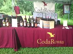 A table with a red tablecloth marked "Coda Rossa" and bottles of wine, which is under a tent with woods in the background.