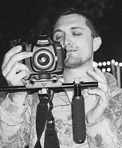 A black-and-white nighttime photograph of a man with a cigarette held in his mouth wearing an autumnal-themed hoodie looking at and adjusting a Canon EOS camera on a rig with microphones attached.