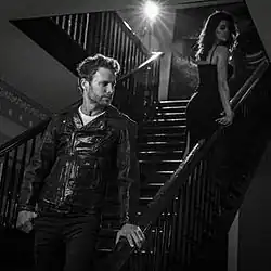 A black-and-white image of Bentley wearing a leather jacket on a staircase, looking at a woman wearing a dress.