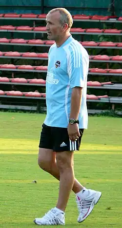 A middle-aged man in a blue shirt is walking on a soccer field.
