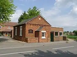 Small red brick building with pitched roof at the roadside on a sunny day against a pale blue and white dappled sky