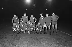 Black and white photo of a football team in kit, with a black sky in the background. Five players are crouched down, and six are standing.
