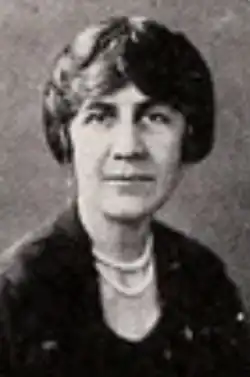 A middle-aged white woman wearing a dark suit and two strands of pearls or beads