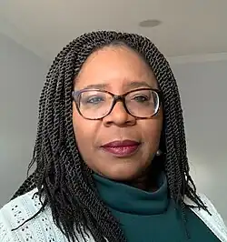 A Black woman with long braids and glasses. Her subtly made up face shows quiet confidence