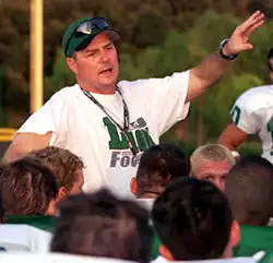 A man in typical football coaching attire, including a hat, large sunglasses, and a lanyard.