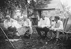 Each of the men sits in a chair outdoors in the grass. Edison reads from a newspaper while the others look at him.