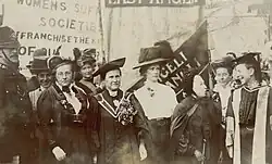 A group of suffragist woman standing together and talking, with banners in the background