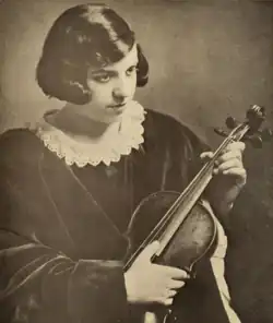 A teenaged girl with fair skin and dark hair, cut in a bob, wearing a dark dress with a lace collar detail; seated, holding a violin