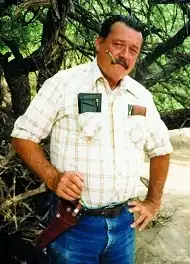 Glenn Boyer standing smoking a cigar with a holstered six-shooter on his hip.