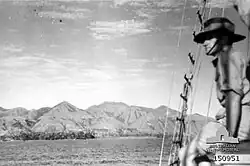 A man in a slouch hat looks out from a sailing ship over a mountainous coastline.