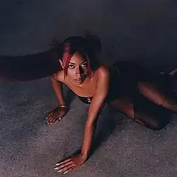Lenae, with red-dyed hair and a sexy black outfit, stares up at the camera while propping herself up with her arms against a mottled floor.