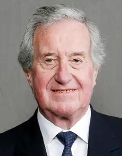 Elderly man with grey hair dressed in a dark blue suit, white dress shirt, and a dark blue-spotted necktie