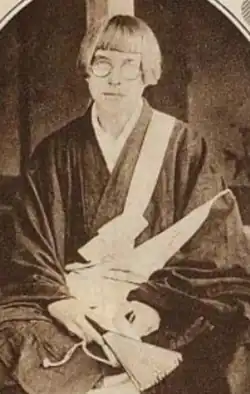 A white woman with blond hair cut in a sharp bob with bangs, wearing round glasses and a dark kimono
