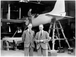 Photograph of two men in suits, in front of an aeroplane, facing the camera.