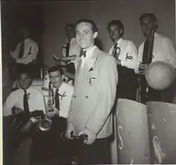 Smiling man standing in front of six other men – all holding musical instruments.