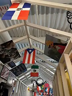 Hallway of La Casita Cultural Center, decorated with hanging flags from the Dominican Republic, México, Puerto Rico, and others. Also features images hanging from the ceiling of important figures like baseball player Ivan Rodriguez, and people from the community like a young girl wearing an orange helmet and a red shirt.