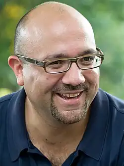 The photo shows a smiling Jeffrey Sánchez from the shoulders up as he looks off to his left. Sánchez is bald, has a goatee-style beard and moustache and is wearing glasses and a dark-blue polo shirt.