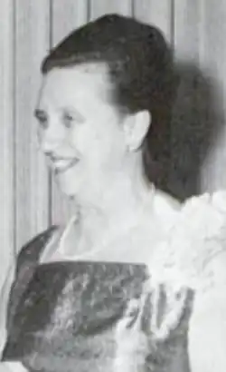 A smiling older white woman, with dark hair in a bouffant updo, wearing a corsage and pearls and a gown with a square neckline