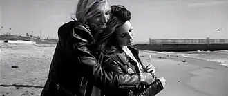 A black-and-white portrait of a brunette woman and long-haired man clinging onto each other while standing on the shore of a beach.