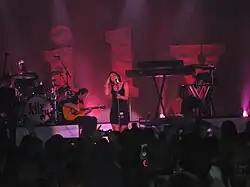 A woman singing on a microphone, standing up, with a band with instruments in her back and a crowd of people in front. There are 3D letters in the background, spelling "Lily".