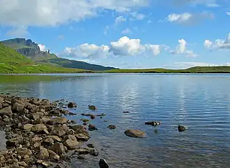A lake, with a stony foreshore and a mountain to the left in the distance
