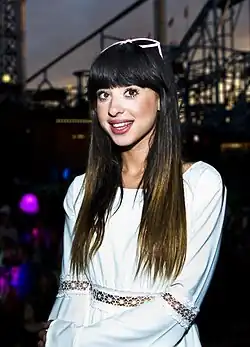 Foxes wears a white dress with lavender sunglasses on top of her head. She stands before a roller coaster and looks at the camera