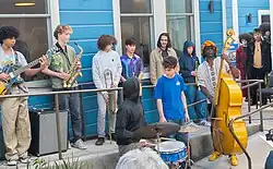 A group of teens play and hold various jazz band instruments while Marcus Shelby plays upright bass. They are standing outdoors next to a blue building with white trim.