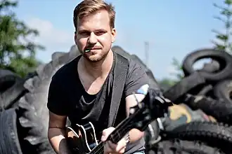 Martin Chodúr wearing a black t-shirt, holding a bass guitar, with a guitar pick in his mouth, standing in a warehouse yard, with various tires in blurred background.