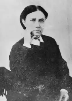 A dark haired woman wearing dark clothing, right hand on chin.