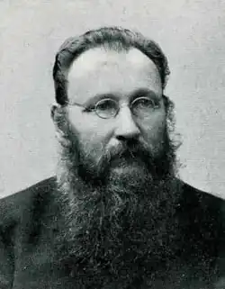 A black and white photograph of a man with a receding hairline, a long beard, and circular lens glasses