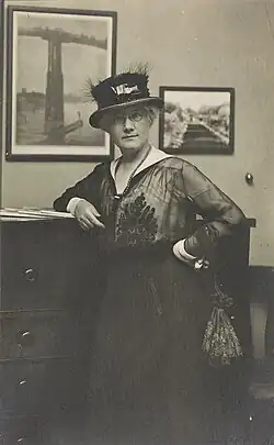 Photo shows Mattie Edwards Hewitt, three-quarter length portrait, standing, facing front, right arm on chest of drawers.