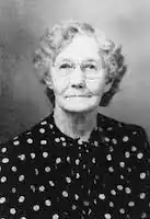 headshot of a white older woman with grey curly hair and glasses wearing a dark shirt with white spots